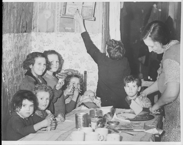 Woman and eight children gathered around a table at meal-time, c. 1935.